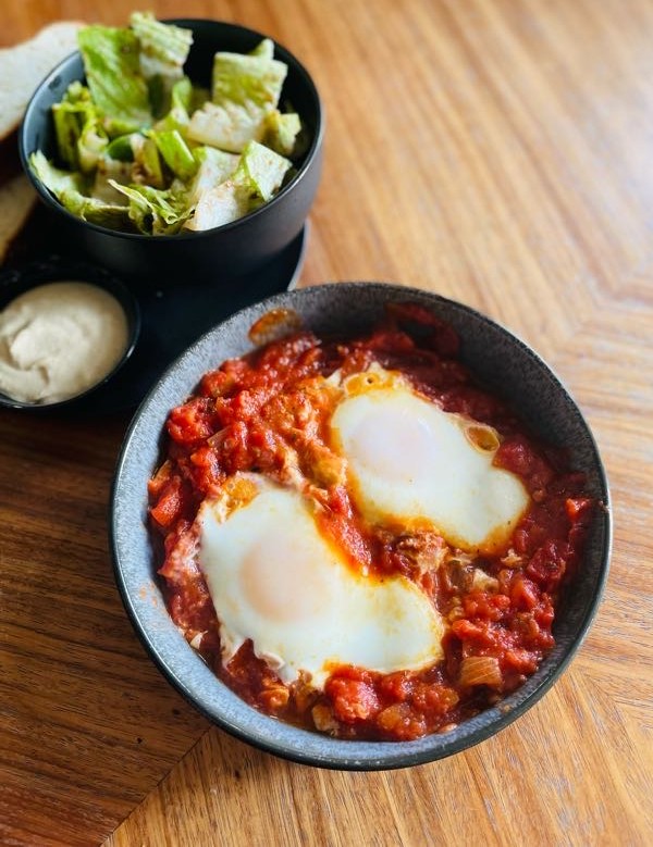 Shakshuka - The Red One Schüssel mit Eiern in Tomatensauce und einer Beilage aus Salat und Dressing.